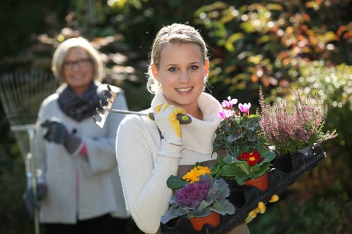 A young woman with fair hair tied back, smiling and wearing a white sweater and gardening gloves, holds a black tray filled with vibrant flowering plants including purple, yellow, and pink blooms, in a well-maintained garden during daylight. Behind her, an older woman with glasses, light-colored hair, and a dark scarf around her neck, also smiling, is holding a garden rake. The garden features a lush green lawn with dense shrubs and trees in the background, displaying a mix of native and flowering plants, and the scene is illuminated by natural sunlight with a slightly blurred background, suggesting a peaceful outdoor space typical of residential gardens in the Hounslow area. This setting exemplifies professional gardening services that enhance outdoor spaces, combining diverse plant varieties, clean paved areas, and well-organised garden layouts. The weather appears clear, contributing to vibrant natural colours and a welcoming atmosphere suitable for gardening projects.