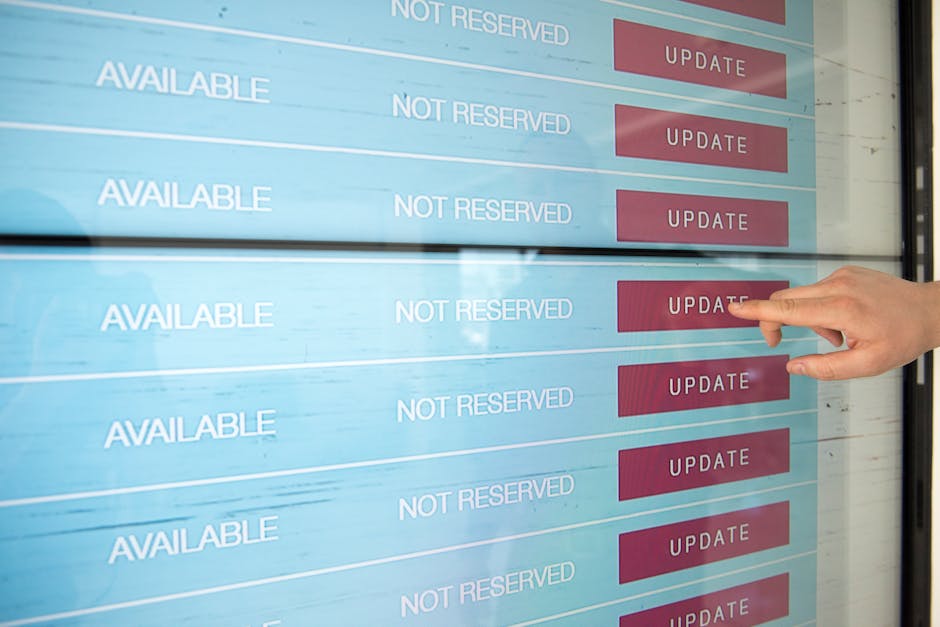 A person's hand pointing at a digital scheduling screen displaying a list of time slots labeled 'AVAILABLE' and 'NOT RESERVED', with red 'UPDATE' buttons beside each slot. The screen is reflected on a